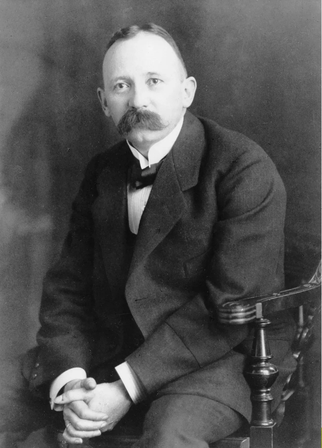 A formal seated studio portrait of a balding man with a heavy moustache, in a dark suit with a bow tie, hands folded in his lap.