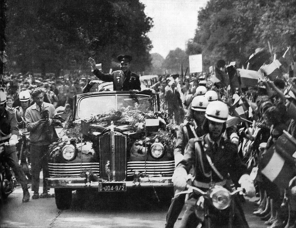 A uniformed man standing in an open-topped car decorated with flowers, waving to a dense crowd lining a tree-lined avenue, with police motorcyclists riding alongside.