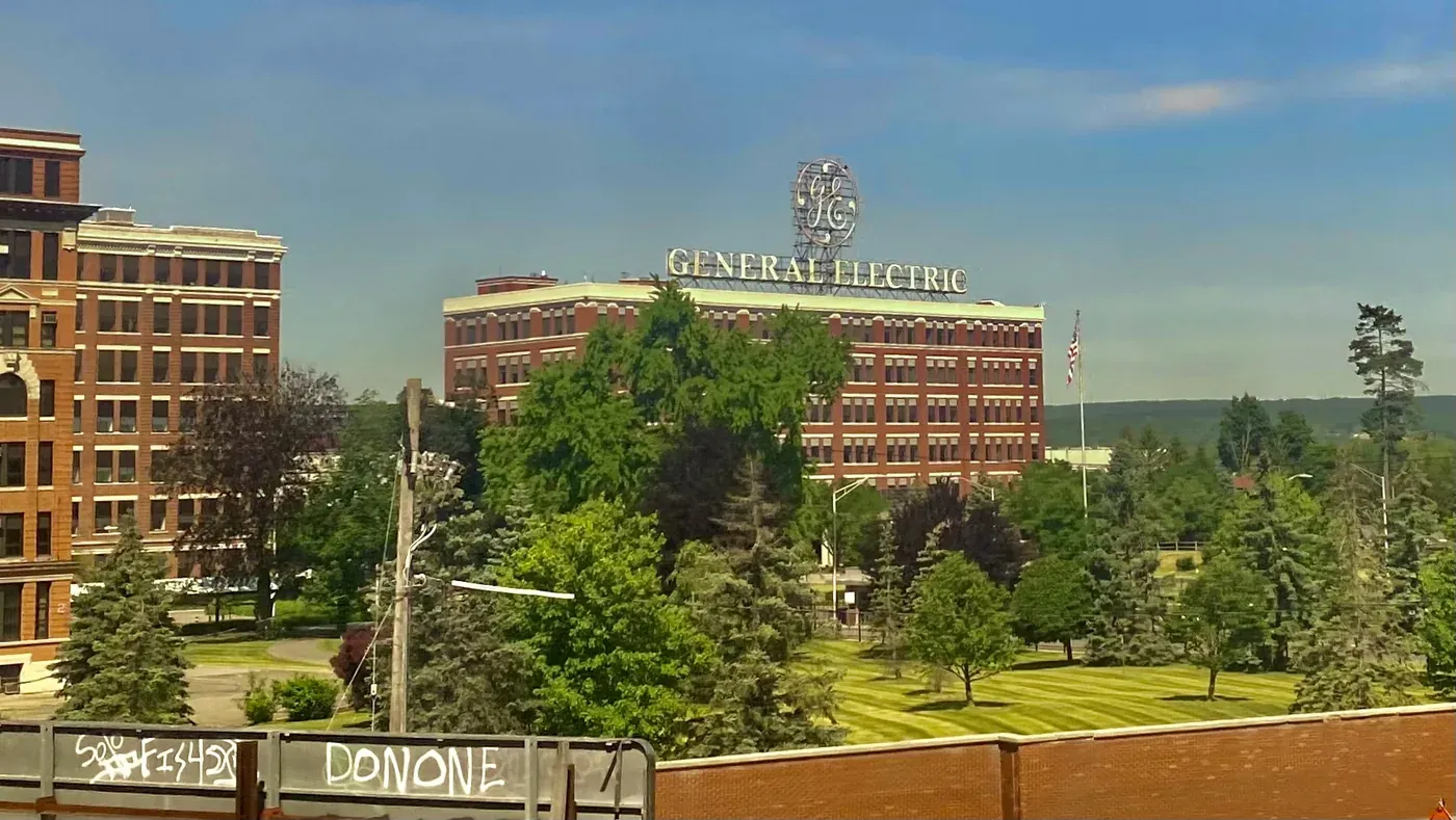 A large early-twentieth-century brick research building with a rooftop sign reading GENERAL ELECTRIC beneath the company's script-lettered monogram, seen across a park-like lawn with mature trees.