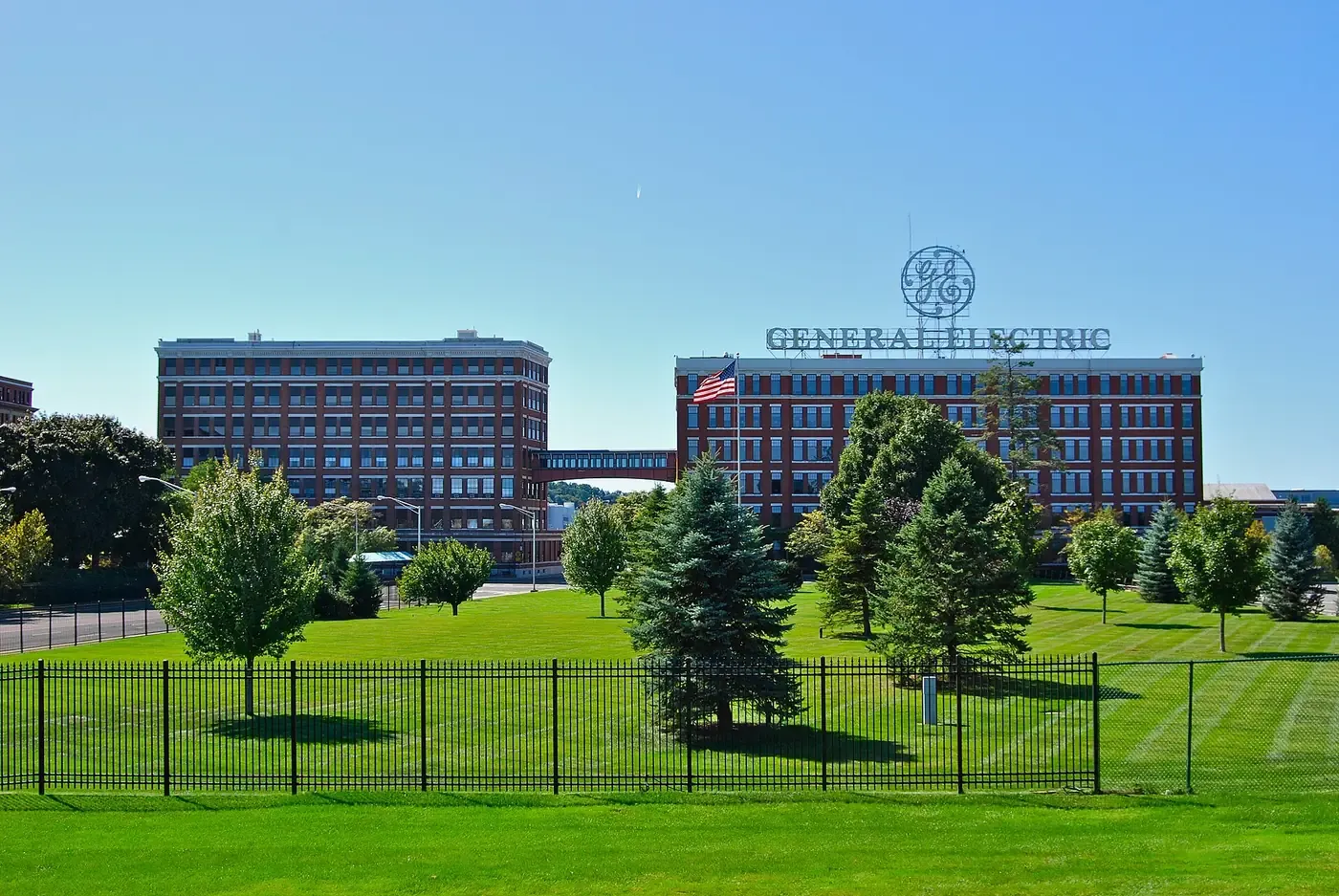 A red-brick early-twentieth-century industrial research complex with a rooftop sign reading GENERAL ELECTRIC beneath the company's monogram, seen across a fenced lawn.