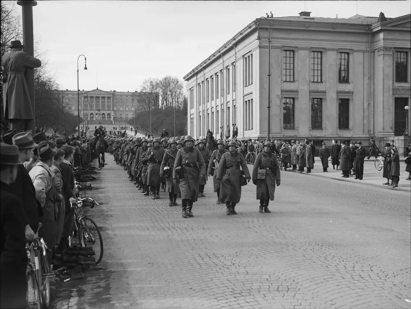 German infantry in steel helmets and long coats marching in formation down a wide cobblestone street past a crowd of Norwegian civilians, bicycles lined against the kerb, with a large stone building beyond them.