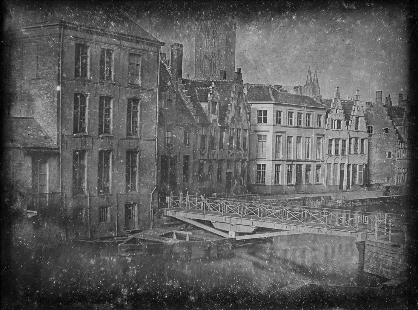 A faded sepia-toned photograph of a canal lined with tall narrow townhouses and a stone bridge, taken from an upper-storey window.