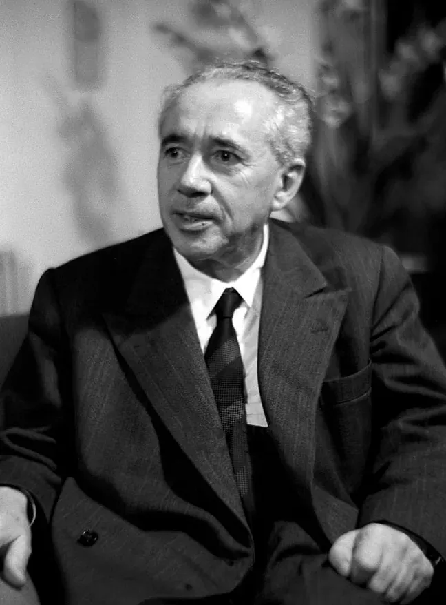A middle-aged man with combed-back grey hair, wearing a suit and tie, photographed mid-sentence in an armchair.
