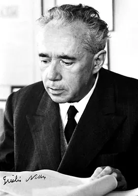 A black-and-white photograph of a man with swept-back grey hair, reading a document at a desk, a signature visible in the corner of the page.