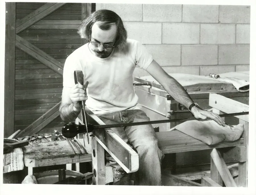 A black-and-white photograph of a man with glasses and a moustache seated at a workbench, shaping a glowing blob of molten glass on the end of a long metal blowpipe resting across wooden supports.