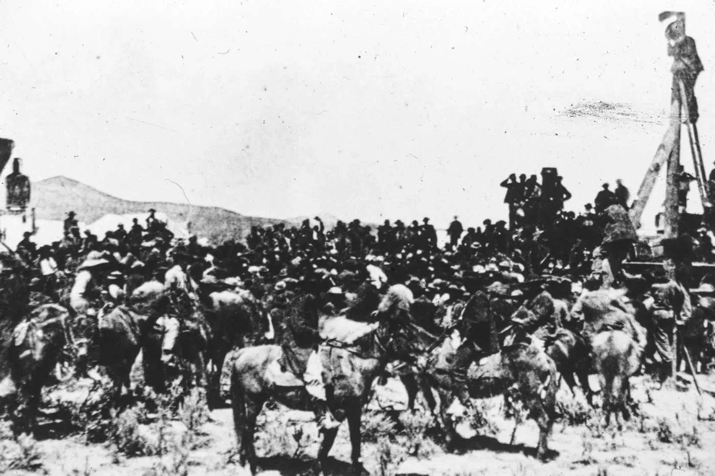 A crowd of men on horseback and on foot gathered around a locomotive and a ceremonial pole at a bare desert railhead, with distant hills behind them.