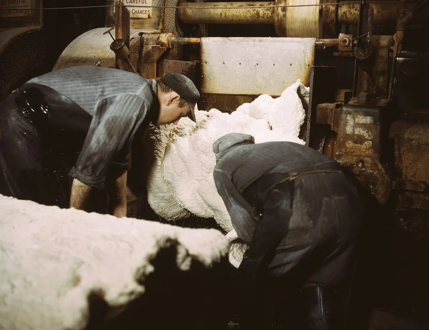 Two workers in overcoats and caps feeding a thick, wet white sheet of rubber into the rollers of an industrial mill, water spraying off the machinery.