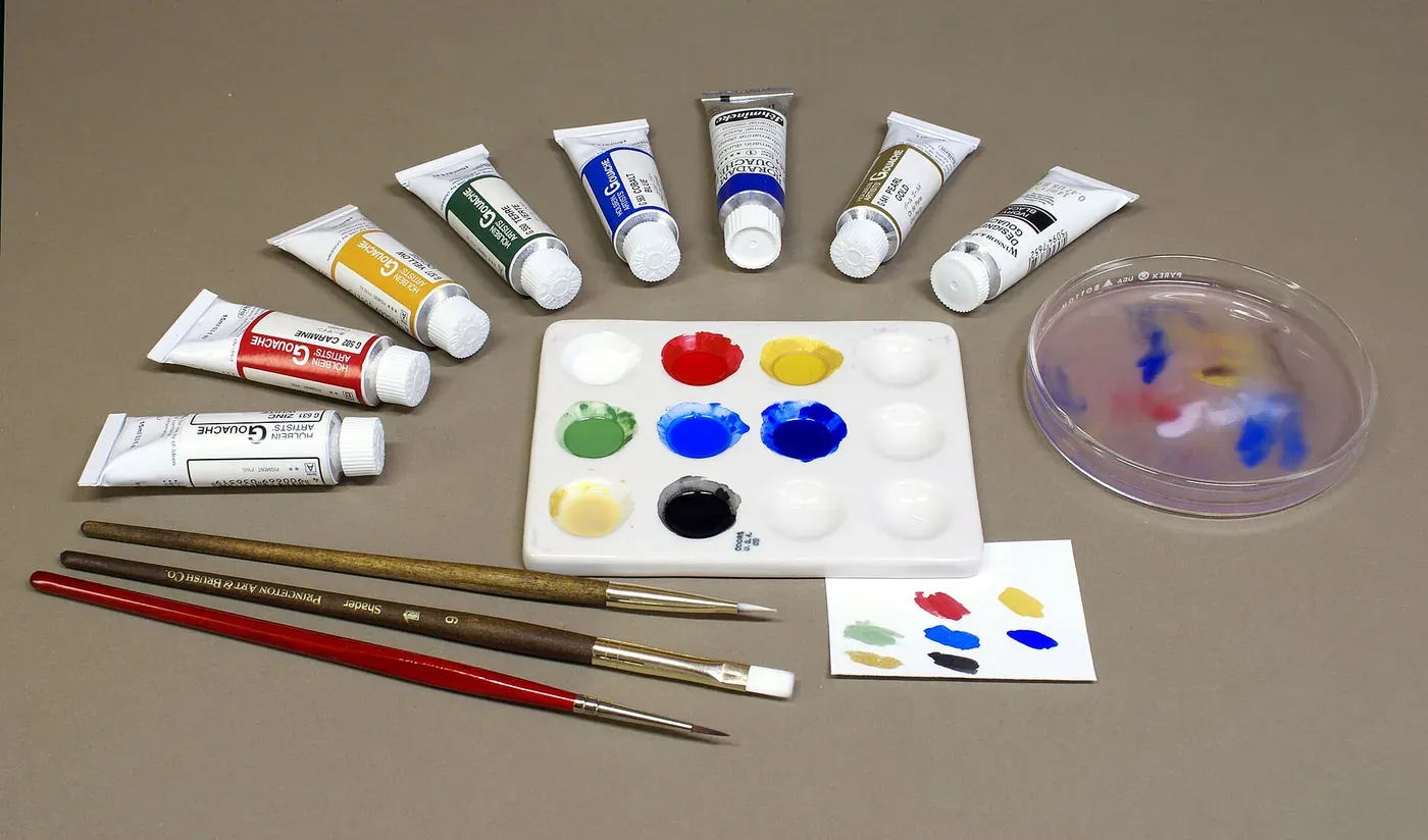 Tubes of gouache paint arranged around a ceramic mixing palette with dabs of red, yellow, blue, green and black paint, alongside two paintbrushes and a water dish.