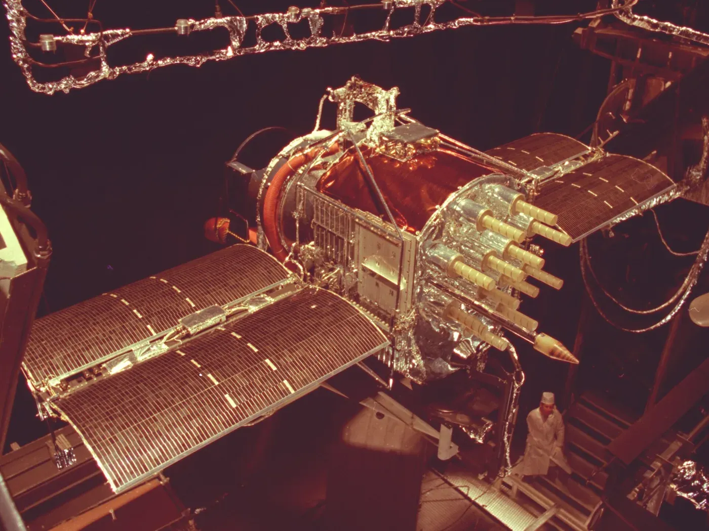 A satellite with a foil-wrapped body and two extended solar panel wings hangs suspended by cables inside a large space-simulation chamber, a technician in white coveralls standing beside it.