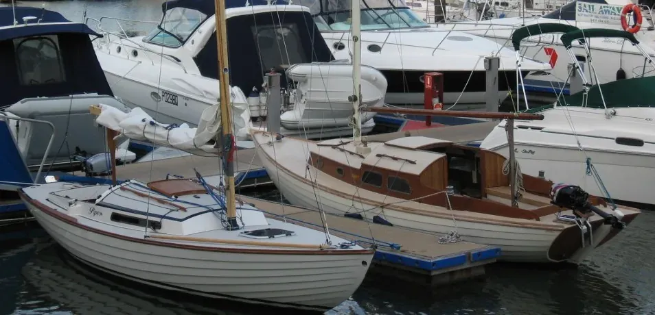A white fiberglass sailboat moored beside an older varnished wooden sailboat of the identical hull design, in a marina with other boats in the background.