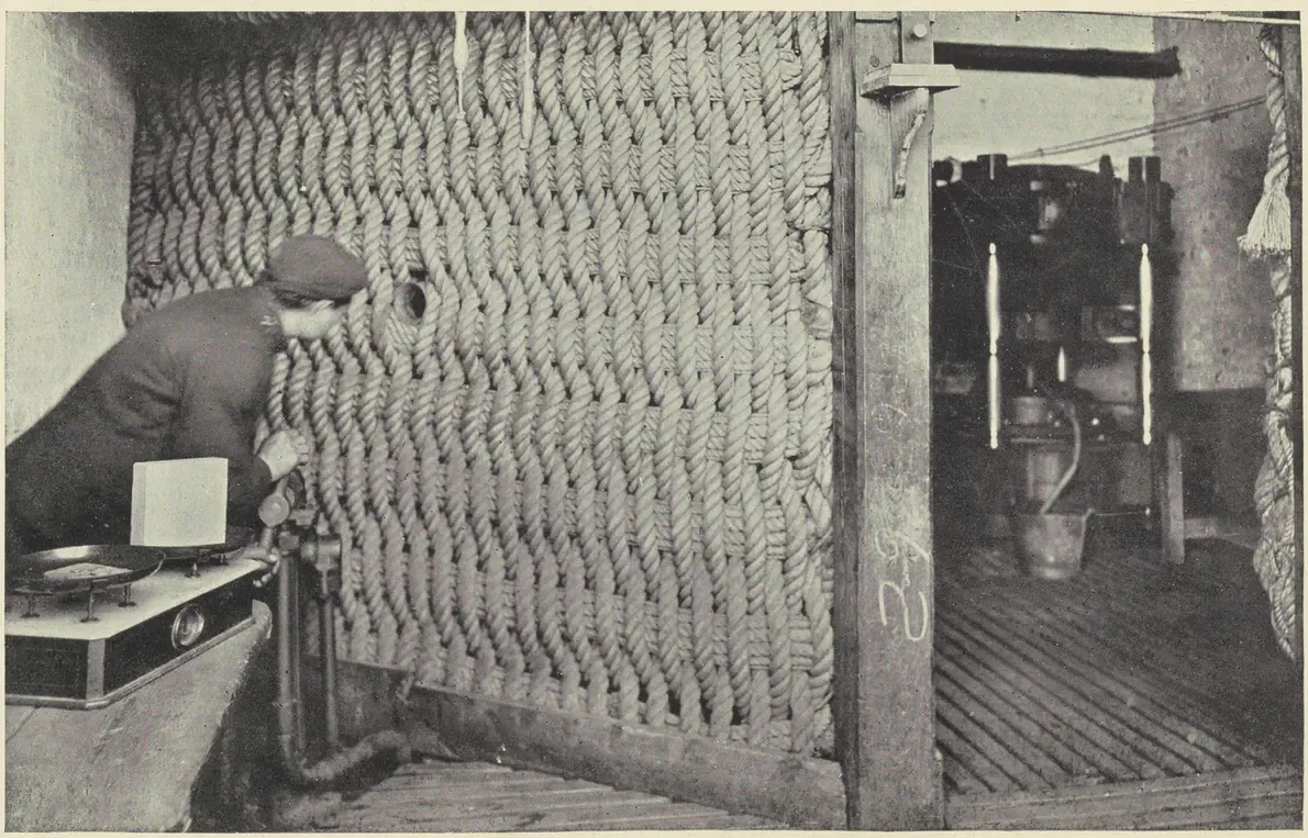 A workman leaning over a thick woven rope screen to reach a guncotton press, with the pressing machinery visible through a doorway beyond the screen.