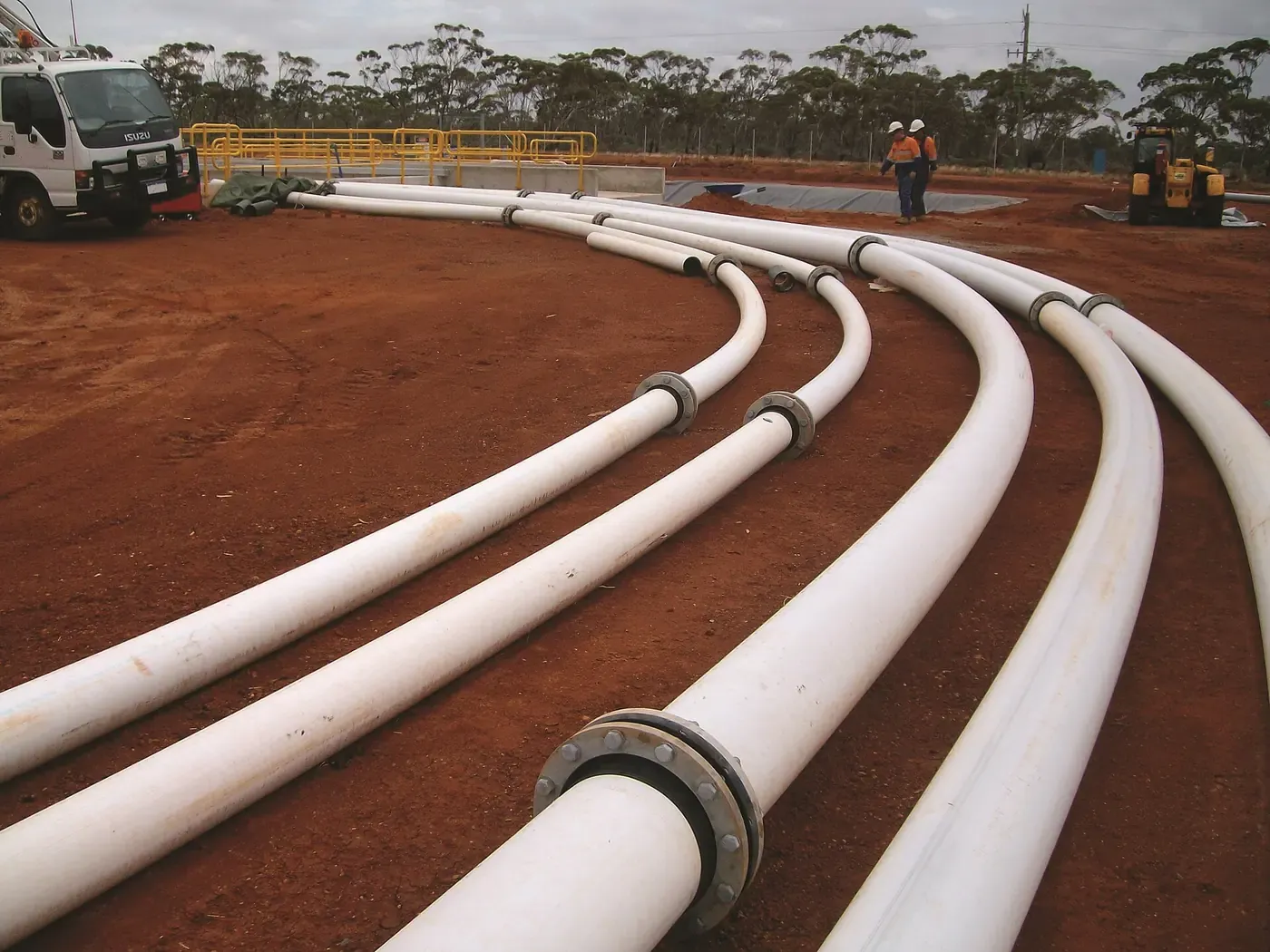 A row of white plastic pipes curving across reddish earth on a construction site, with workers in high-visibility clothing and machinery in the background.