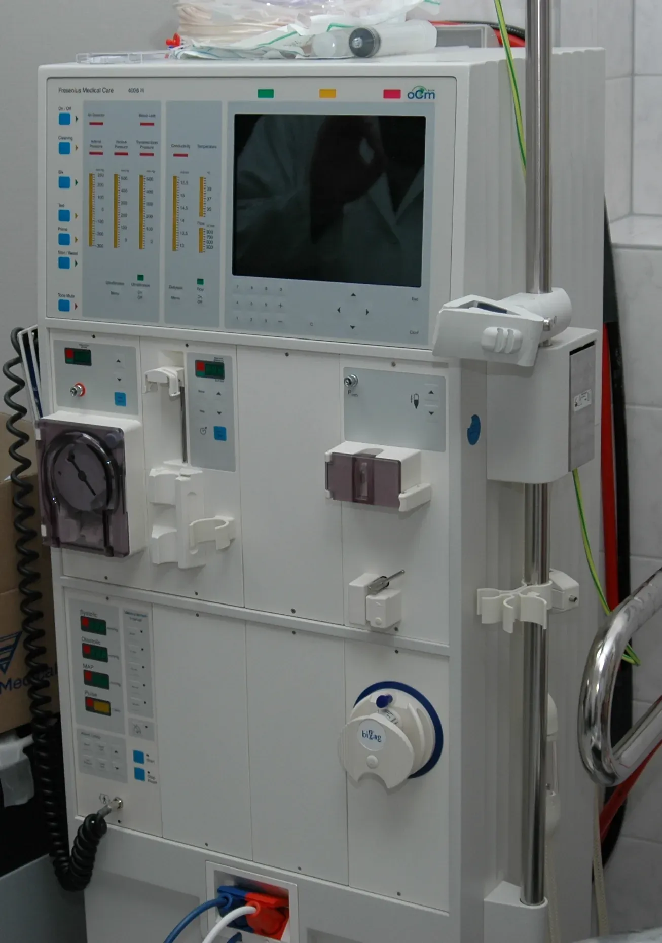 A tall white hemodialysis machine with a control panel of dials, buttons and a small screen, alongside pump mechanisms and tubing, in a tiled clinical room.
