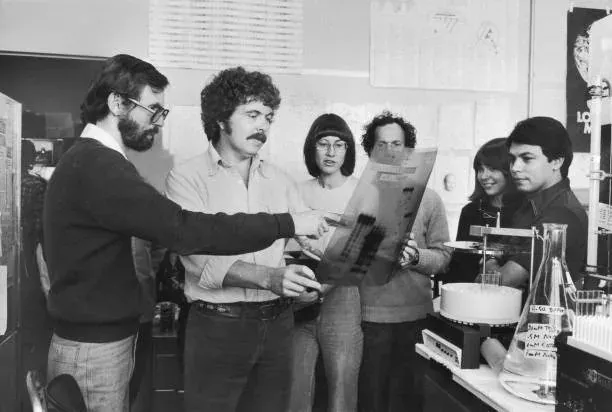 A black-and-white photograph of six researchers crowded around a laboratory bench, one bearded man holding up a large striped autoradiograph film while colleagues look on, flasks and hand-labelled bottles visible on the bench to the right.