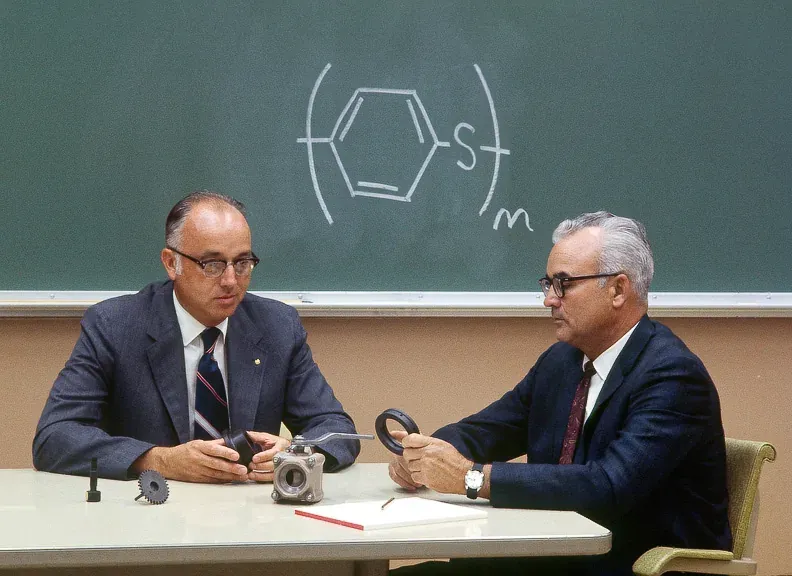 Two men in suits sit at a table examining a molded gray plastic valve component and a small gear, in front of a chalkboard chemical structure of a benzene ring bonded to sulfur, labeled with a repeating-unit bracket.