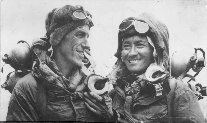 Two mountaineers in cold-weather climbing gear and oxygen masks smiling at each other, oxygen cylinders and equipment visible on their backs.