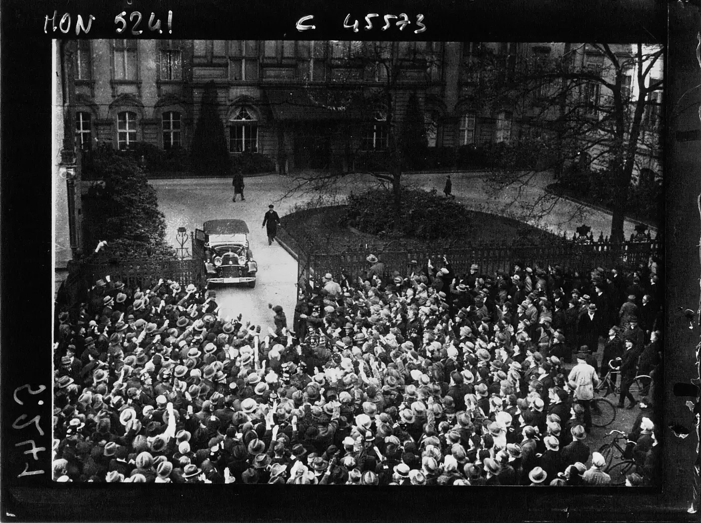 An open-topped car creeping through a dense crowd toward a palace entrance, with people pressed against iron railings on both sides waving hats and papers, photographed from an upper window.