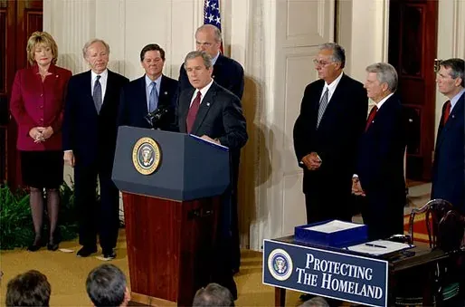 President George W. Bush speaking at a podium marked 'Protecting the Homeland,' flanked by a row of officials, inside a formal room with an American flag.