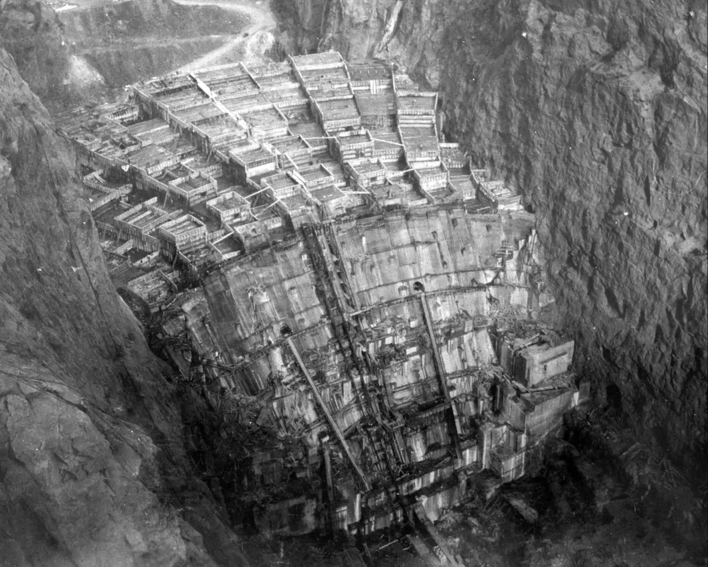 An aerial view down into a steep rock canyon showing the top of a dam under construction as a grid of individual rectangular concrete columns, each poured separately, still open at the top, with cranes and pipework running between them.