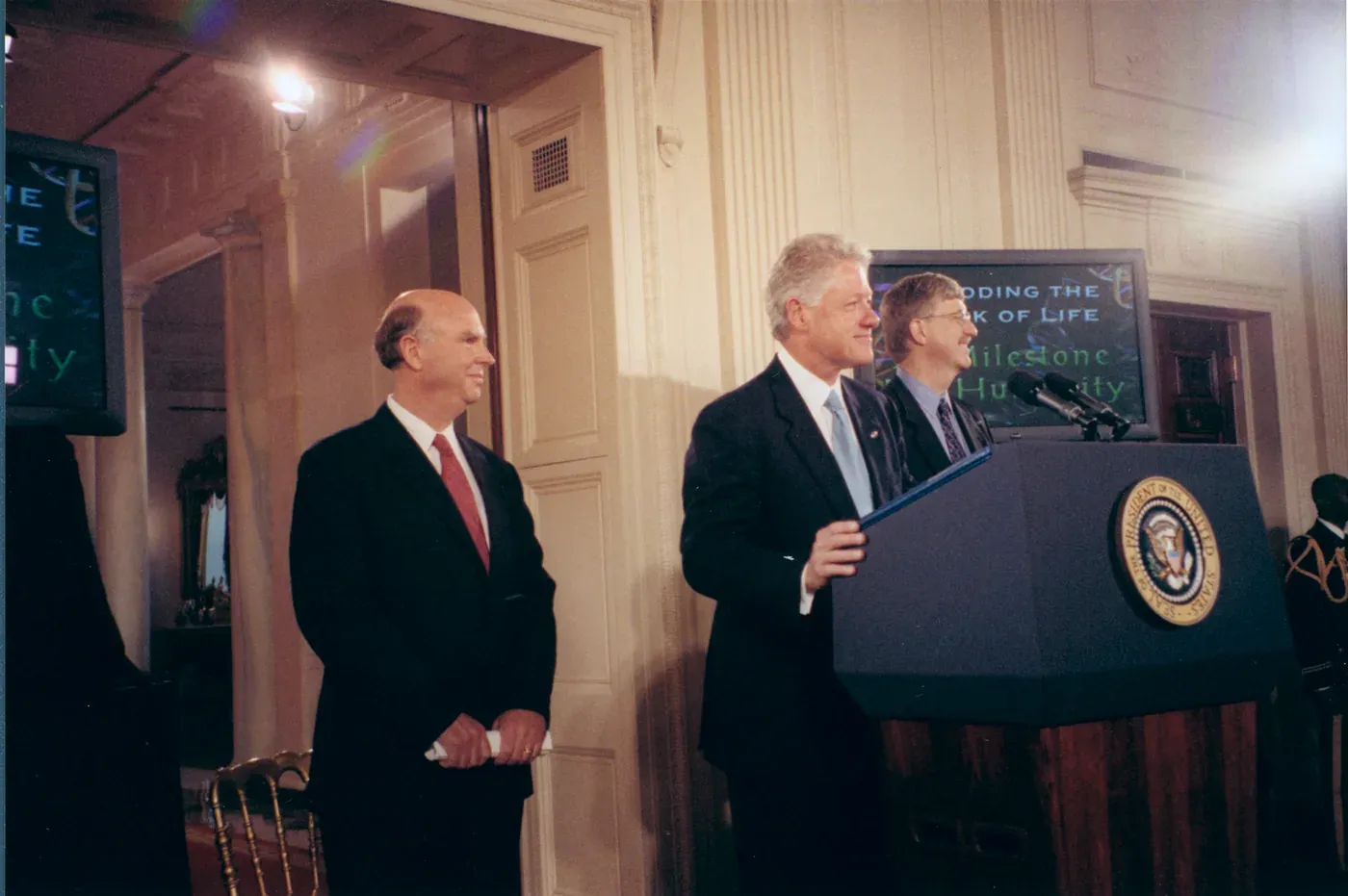 Bill Clinton speaking at a lectern bearing the Presidential seal, flanked by two other men, with screens behind them reading a headline about decoding the book of life.