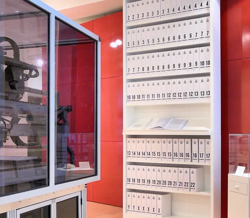 A tall white bookcase holding more than a hundred identical spine-labelled volumes, each printed with a run of chromosome numbers, standing against a red wall in a museum gallery.