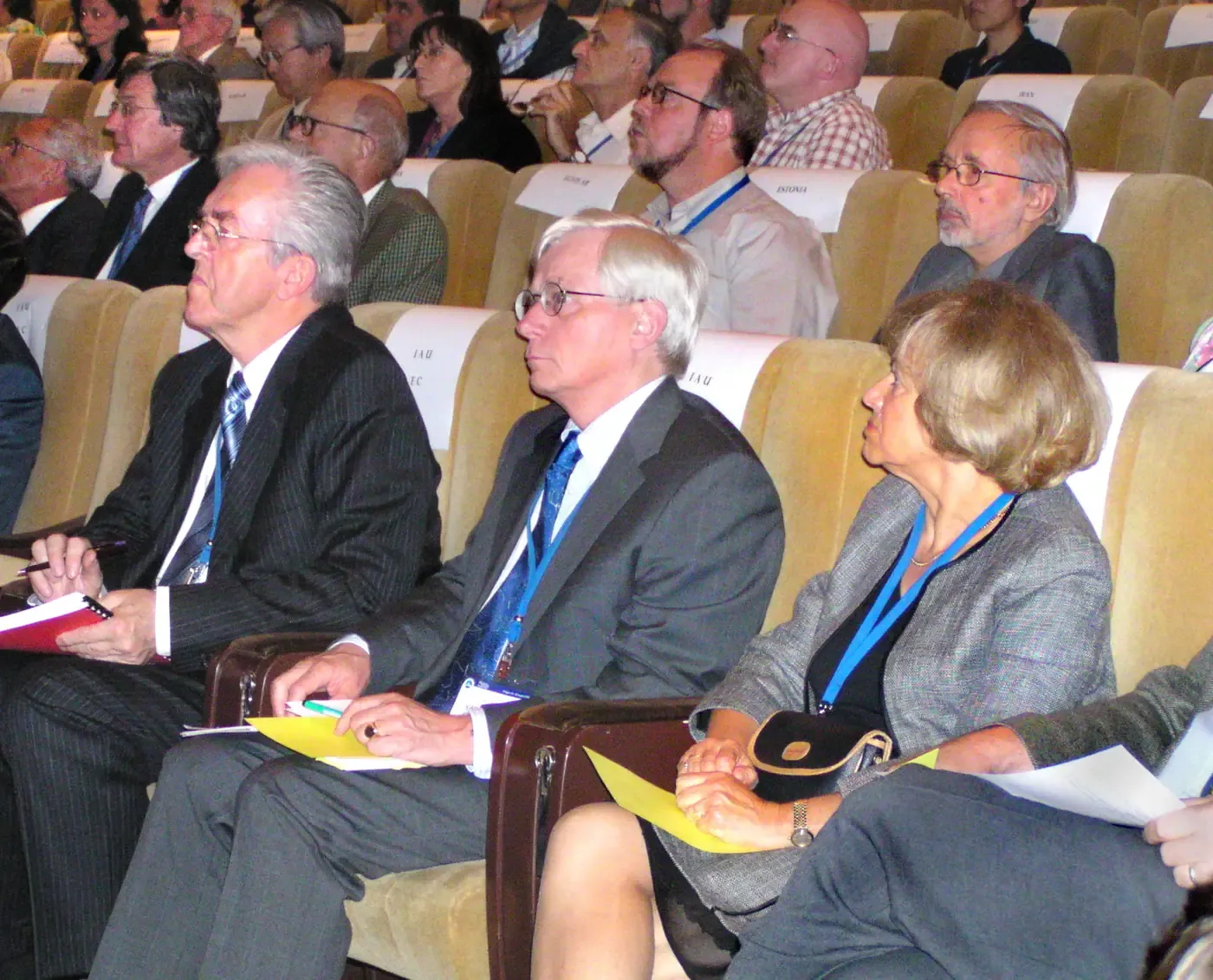 Rows of seated delegates in a theatre-style hall, several wearing lanyards and holding papers, listening attentively during a conference session.
