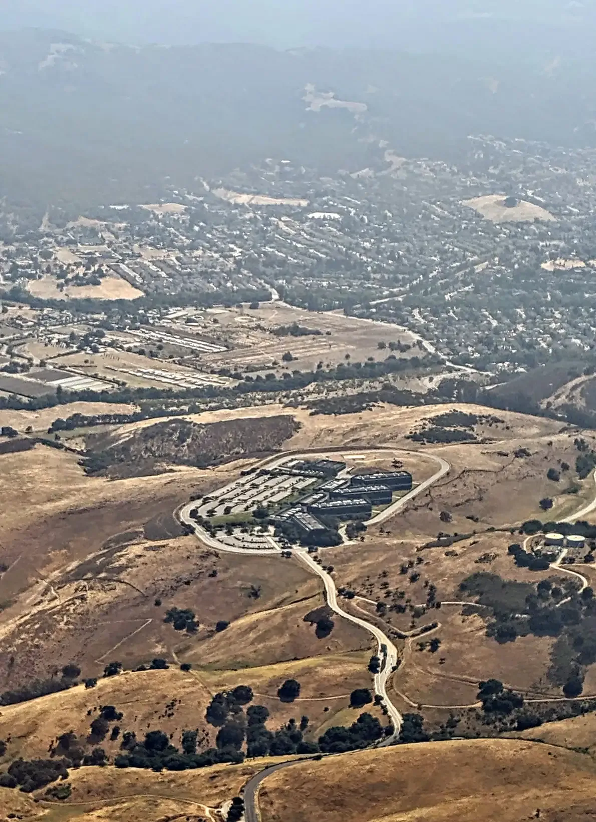 An aerial view of a low, dark, multi-winged office building set among dry golden hills, reached by a single winding access road, with a small town visible in the hazy valley beyond.