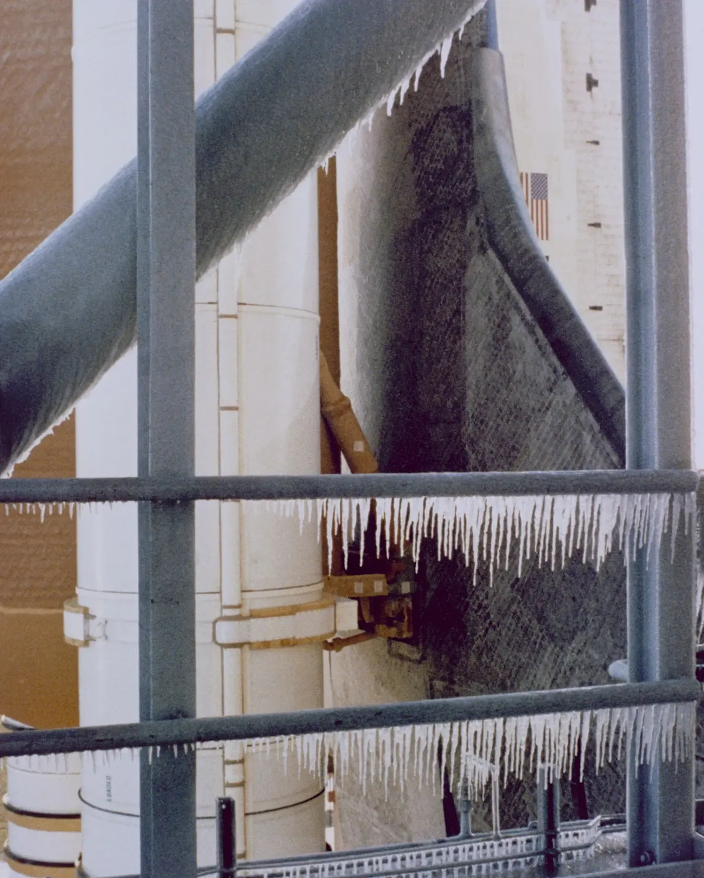 Thick icicles hanging from a metal launch-tower railing and structural beams, with the frost-covered white flank of a Space Shuttle stack visible behind.
