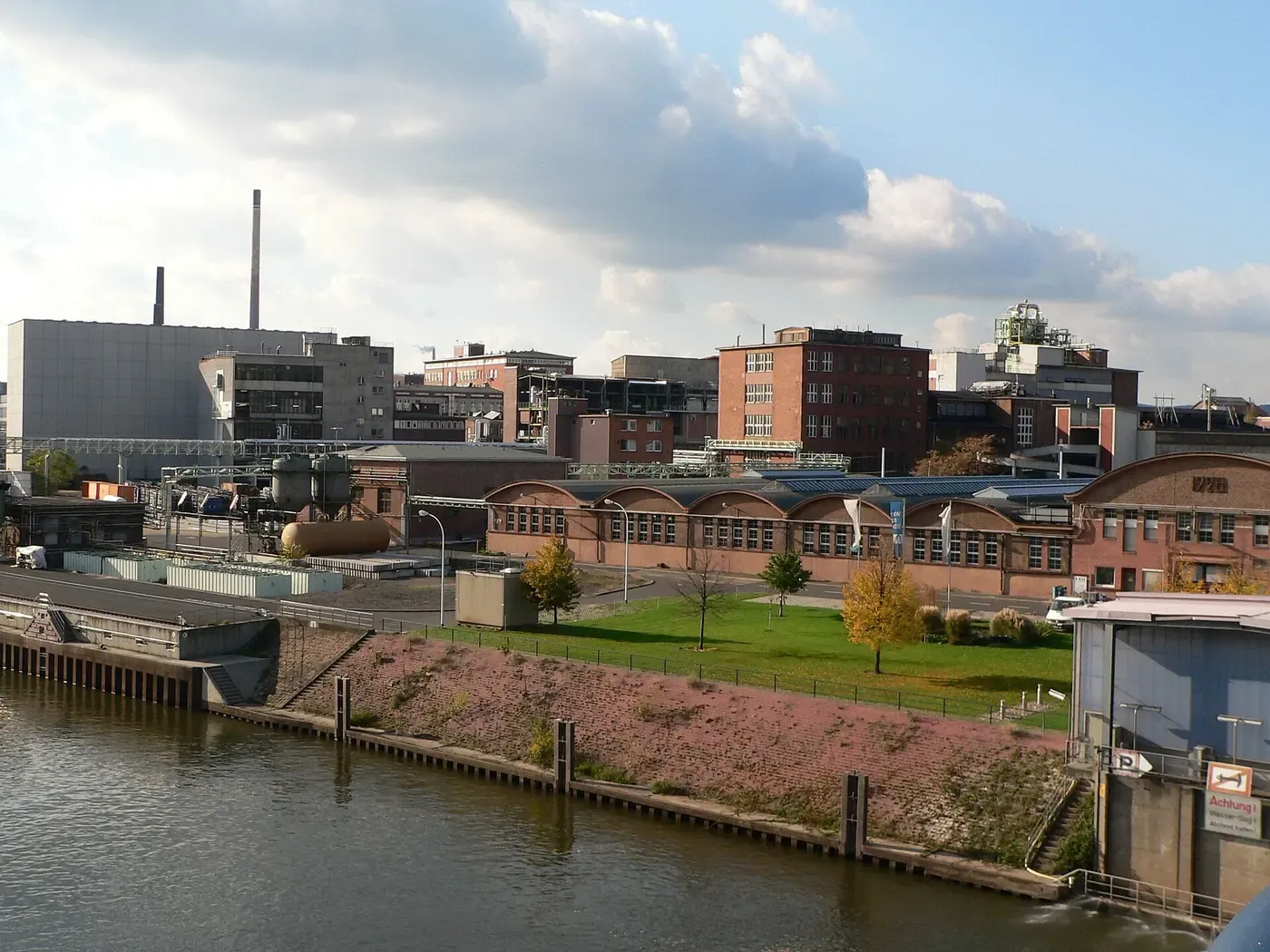 A riverside industrial site with brick factory buildings, a tall chimney and pipework, seen across the water from a bridge.