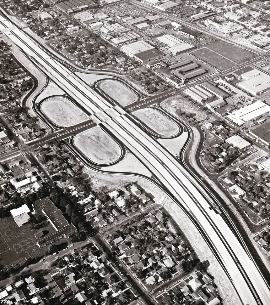 Aerial black-and-white photograph of a newly built cloverleaf highway interchange cutting through a grid of suburban houses and small factories.