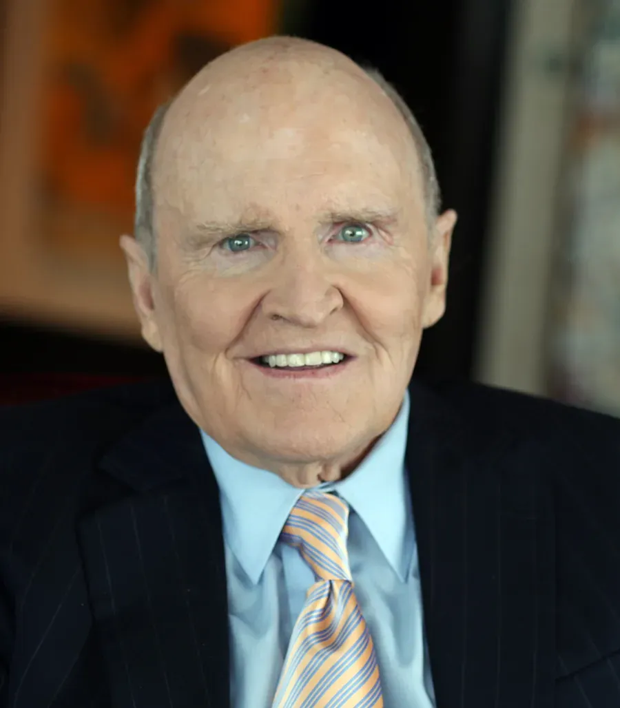 A bald, smiling older man in a dark pinstriped suit, light blue shirt and striped tie, photographed head-on in a portrait setting.