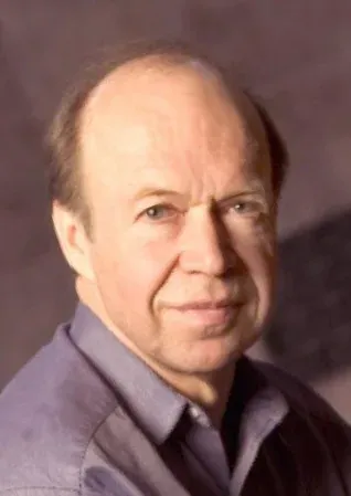 A head-and-shoulders portrait of a balding man with grey hair, wearing a purple button-down shirt, looking directly at the camera.