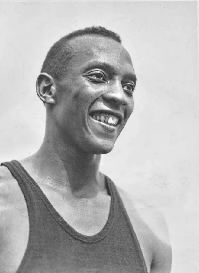 A close head-and-shoulders photograph of Jesse Owens, laughing, in a dark athletic vest, his head shaved.