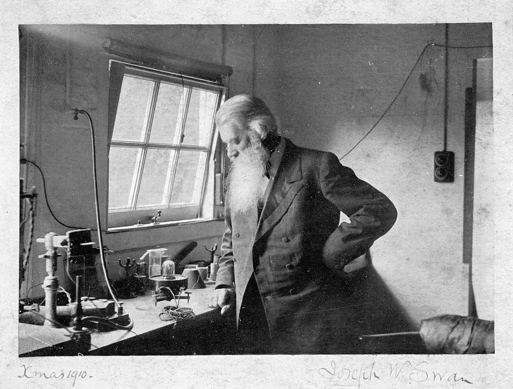 An elderly, long-bearded man in a dark coat leans on a laboratory bench cluttered with glass apparatus beside a window, signed 'Joseph W. Swan, Xmas 1910.'