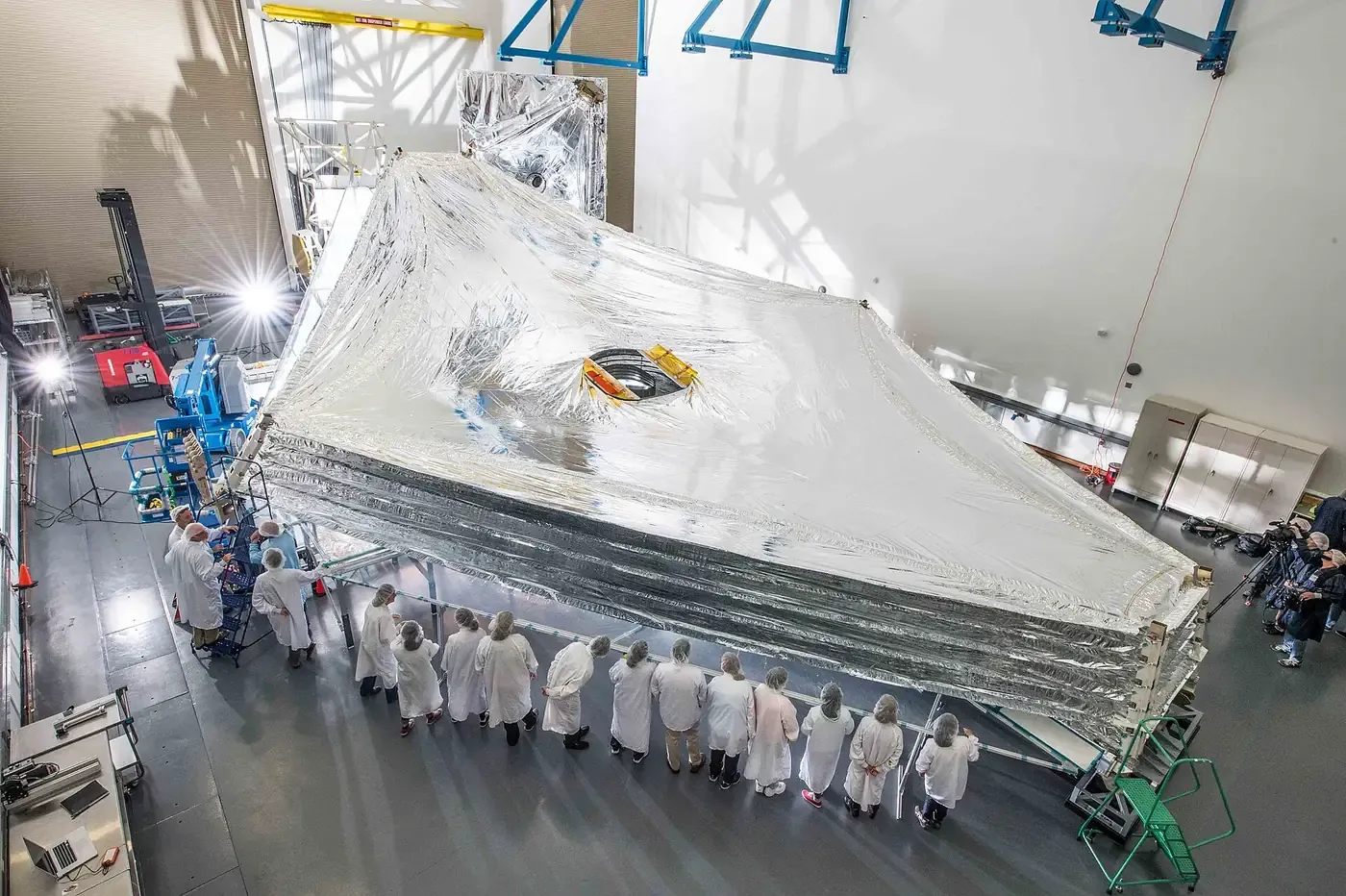 A large, tennis-court-sized metallic silver membrane, folded into a tent-like shape, being inspected by a row of technicians in white cleanroom suits inside a high-bay facility.
