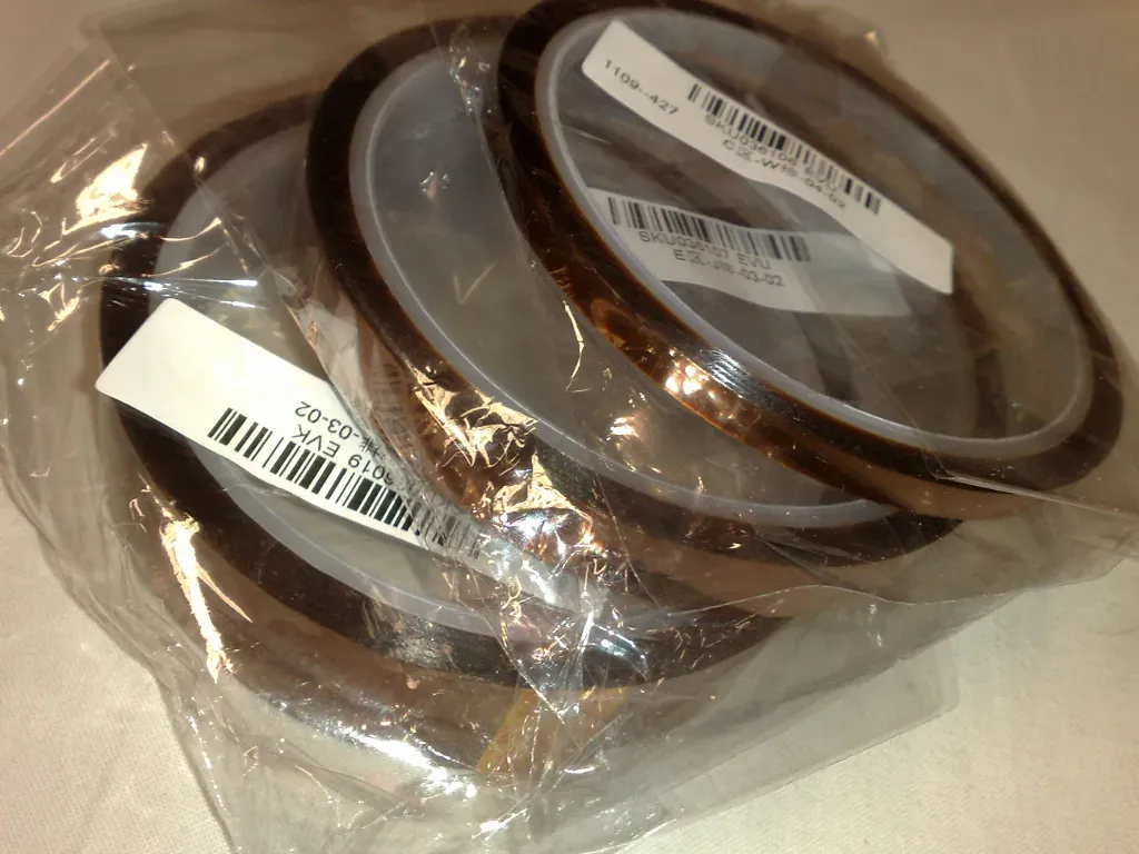 Several rolls of amber, semi-transparent Kapton polyimide tape, still shrink-wrapped with barcode labels, stacked together.