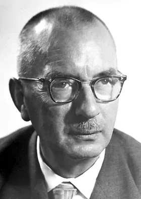 A black-and-white portrait photograph of a balding man wearing thick horn-rimmed glasses and a dark suit, looking directly at the camera.