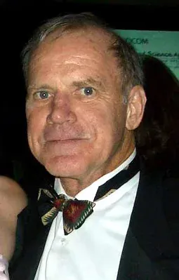 An older man with grey hair, wearing a black tuxedo jacket and a patterned bow tie, photographed at a formal event.