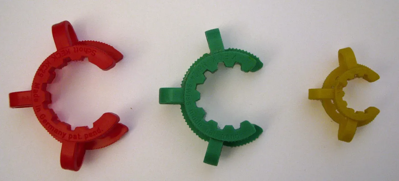 Three plastic Keck clips shaped like the letter C with a locking tooth, in red, green and yellow, of decreasing size, lying on a white surface, each stamped with a size code and 'Made in Germany' or 'Swiss made'.