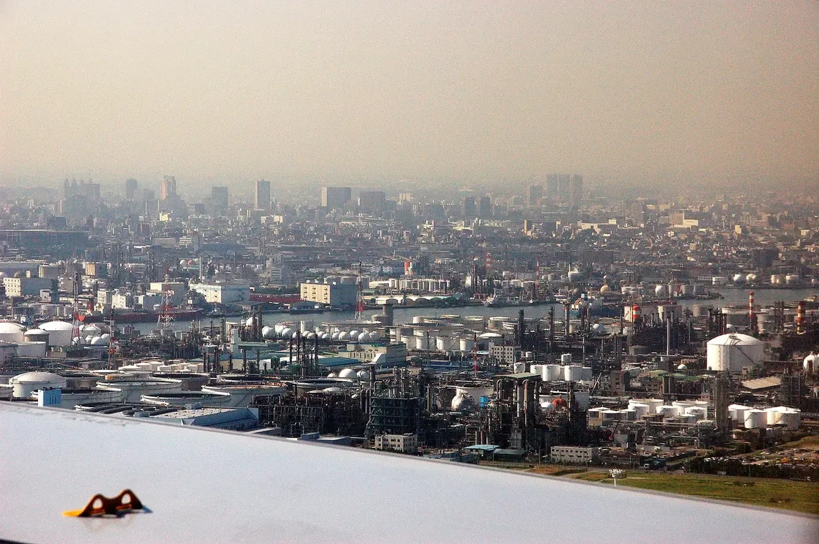 An aerial view over a dense industrial waterfront, packed with storage tanks, chimneys, and refinery towers, with a hazy city skyline visible in the distance beyond.