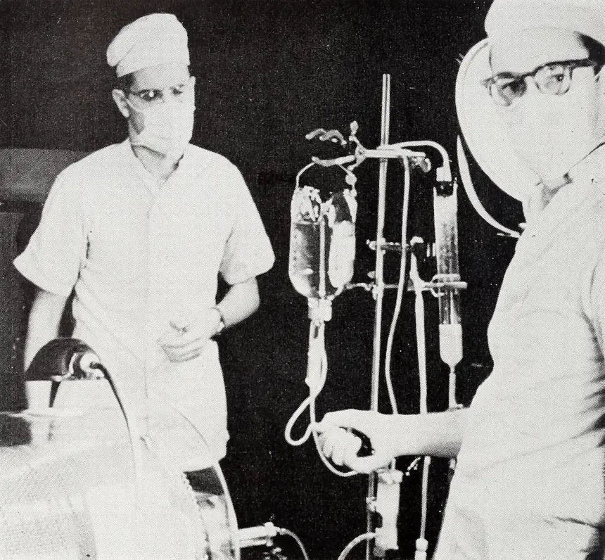 Two masked, capped medical staff in a hospital ward standing beside a stand of glass bottles and rubber tubing connected to an artificial kidney machine, partially visible at the lower edge of the frame.