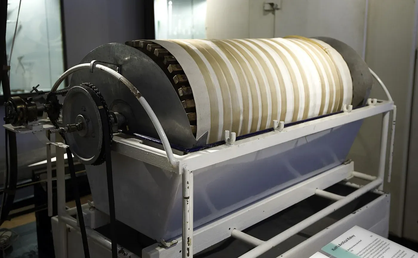 A preserved rotating-drum dialysis machine on display in a museum: a wide horizontal drum wound with cellophane tubing, mounted in a white metal cradle over a shallow trough, with a hand crank and drive chain to one side.