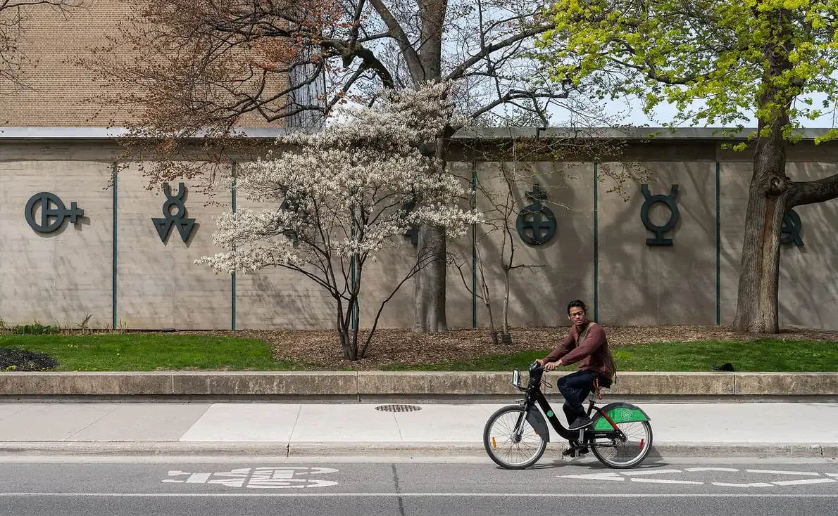 A low, symbol-decorated concrete wall along a University of Toronto street, part of the chemistry building, with trees in blossom and a cyclist passing on the sidewalk.