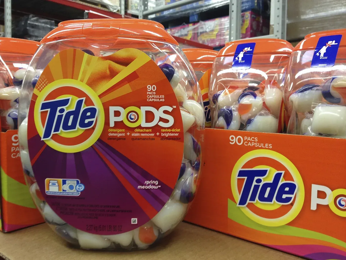 Clear plastic tubs of Tide Pods laundry detergent capsules on a store shelf, each capsule visible as a multicolored gel unit wrapped in a thin transparent film.