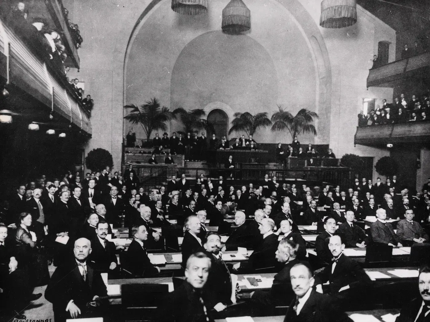 A large domed assembly hall packed with rows of delegates in dark suits seated at desks, potted palms flanking a raised dais at the back, with spectators crowded into balconies on either side.
