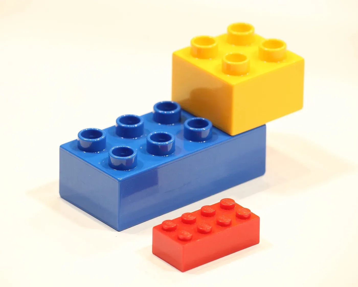 Three plastic interlocking building blocks of different sizes (a small red brick and two larger blue and yellow blocks with rounded studs on top) on a plain light background.