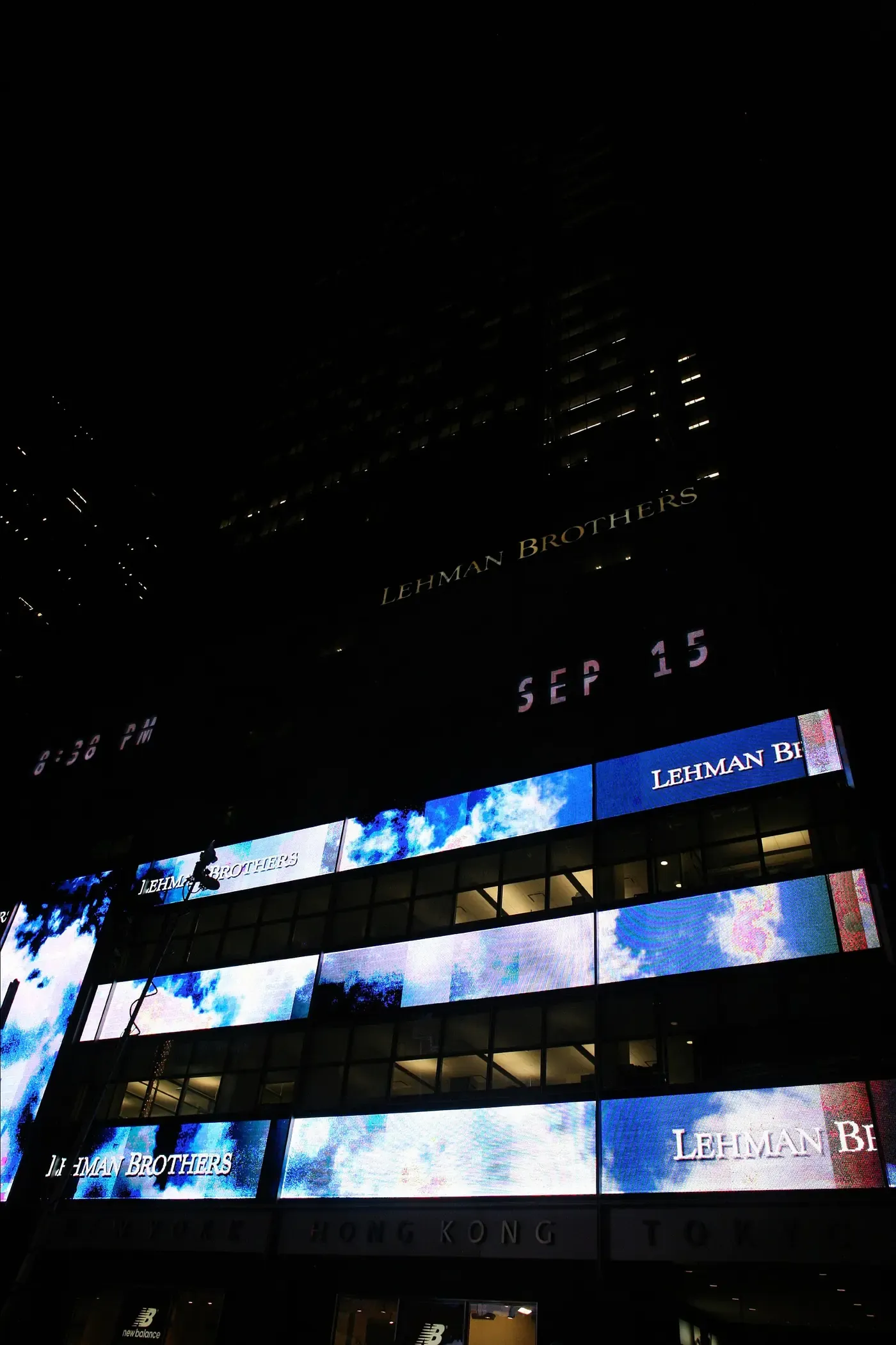 A dark New York skyscraper at night, its lower floors covered in illuminated electronic screens reading 'LEHMAN BROTHERS' and the date 'SEP 15', photographed from street level looking up.