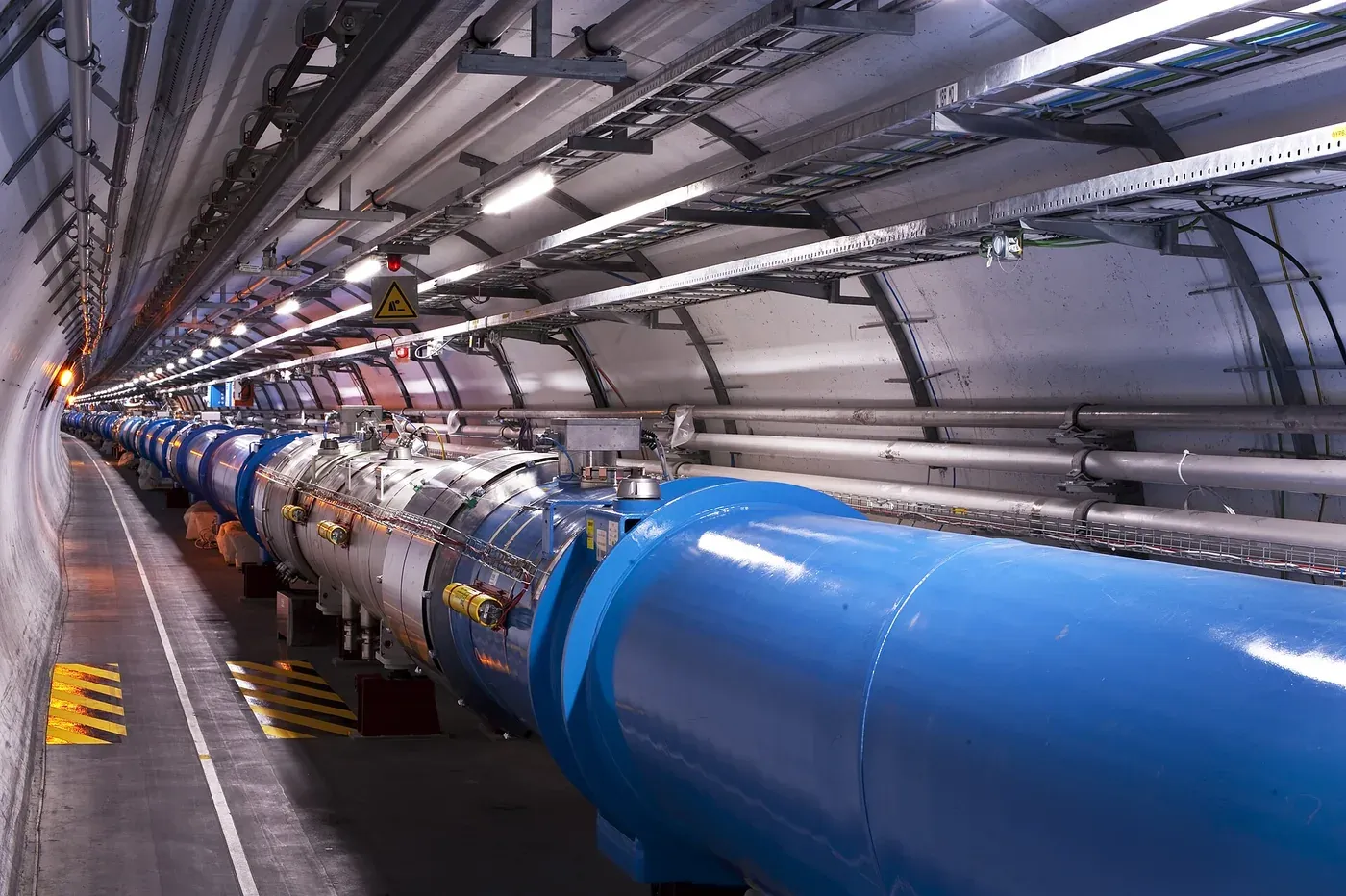 The curved interior of an underground tunnel lined with large blue and silver cylindrical magnet segments running into the distance, overhead cable trays and warning signage visible along the ceiling.