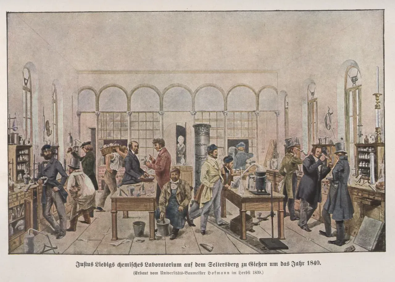 A hand-colored illustration of a long, high-windowed chemistry laboratory around 1840, crowded with students in coats and top hats working at benches lined with glassware and furnaces.