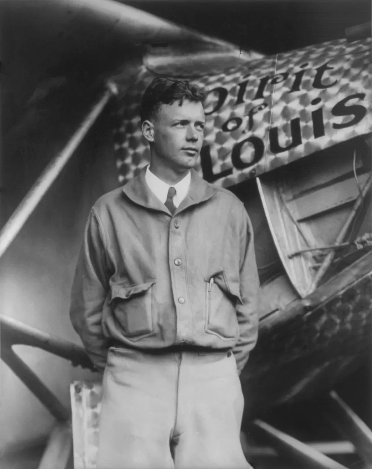 Charles Lindbergh standing in flight jacket and tie in front of the nose of the Spirit of St Louis, the words 'Spirit of' and 'Louis' visible on the fuselage.